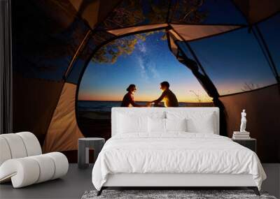 View from inside tourist tent. Young romantic couple, man and woman sitting at campfire holding hands on sea shore on evening starry sky at sunset and sea water background. Tourism and love concept. Wall mural