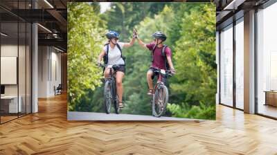 Young couple ride on a mountain asphalt road in the woods on bikes with helmets giving each other a high five Wall mural