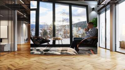 Young man enjoying weekends inside contemporary barn house. Happy male tourist sitting on chair, holding cup of tea, enjoying view of mountain landscape through panoramic windows in new cottage. Wall mural