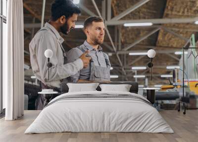 A male inspector or operator of a workshop for the production of aluminum and plastic wreaths trains an intern. International team of men working together near a machine in a factory. Wall mural