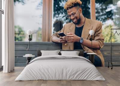 A portrait of African American man in jacket and glasses with smartphone sitting near the windows Wall mural