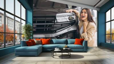 Charming young Caucasian woman hugging hood of new car and show fist and say yes in modern bright car dealership. Happy girl buyer rejoices after making her dream come true. Wall mural