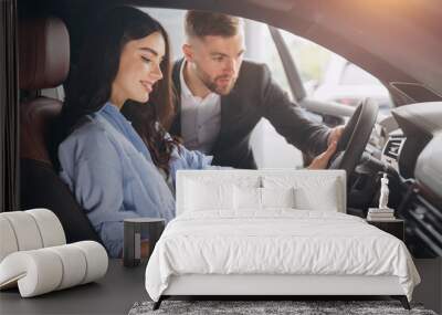 Friendly male manager showing young female customer cars. Beautiful smiling woman sitting inside vehicle in car. Bearded sales consultant working in car dealership. Wall mural