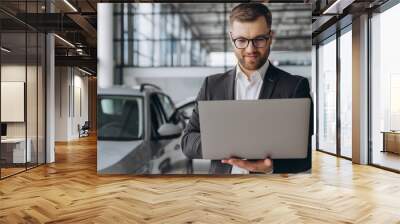 Modern bearded man in glasses and suit vehicle sales consultant using laptop inside car dealership Wall mural