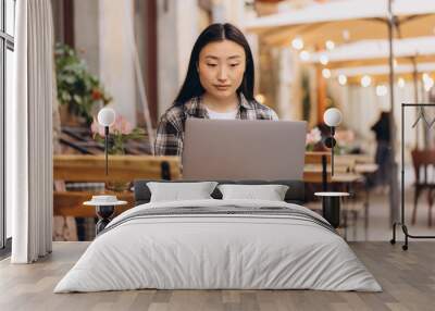 Portrait of a beautiful brunette Korean freelancer sitting in an authentic cafe and using a laptop computer and working on it. Wall mural