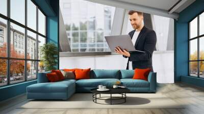 Portrait of handsome happy businessman man in suit holding laptop on background of urban buildings and offices. Wall mural