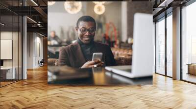 Portrait of happy african businessman using phone while working on laptop in a restaurant. Wall mural