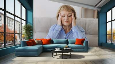 Portrait of senior woman sitting on a sofa at home with a headache, feeling pain and with an expression of being unwell Wall mural