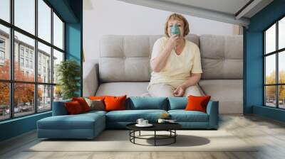 Sick senior woman has a viral infection or flu, labored breathing, asthma. An elderly female is using inhaler, nebulizer for self treatment sitting alone on the sofa at home Wall mural