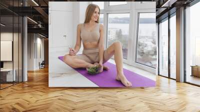 Sporty and pretty blonde woman in sportswear sits on yoga mat in her apartment and eats fresh vegetable salad after workout. Wall mural