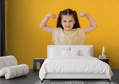 Strong powerful beautiful caucasian little girl over yellow background toothy smile, raises arms and shows biceps Wall mural