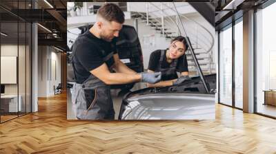 Trying to identify the source of the problem. Two mechanics man and woman working together at open hood on a car in an auto repair shop. Wall mural