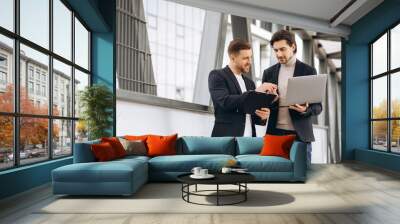 Two handsome young male businessmen in suits hold a laptop and a folder with documents and discuss a working project or have a video call against the background of urban office buildings. Wall mural