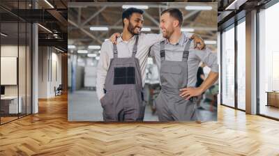 Two happy international workers in overalls hug each other against a factory background Wall mural