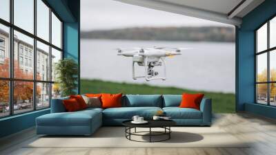 White quadrocopter is flying high in the air, taking photos, recording footage from above. Drone with four motors and propellers, camera and red warning lights. Wall mural