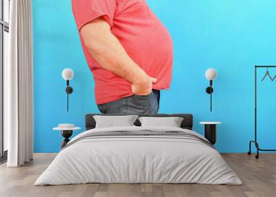 A fat man with a big belly in jeans and a red T-shirt on a blue background holds his hands in his pockets. Wall mural