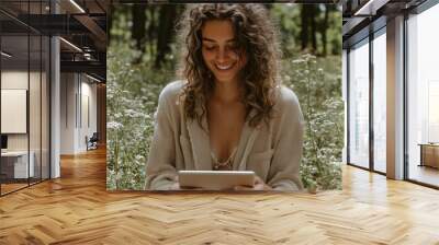 Young woman with curly hair smiles while using a digital tablet, sitting in a forest clearing surrounded by white flowers, enjoying technology in nature Wall mural