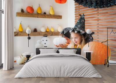 Cute little girl in witch costume sitting behind a table in Halloween theme decorated room, holding hand painted pumpkins and smiling. Halloween holiday concept. Wall mural