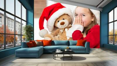 Cute young girl wearing santa hat whispering a secret to her teddy bear christmas present toy. Cheeky kid sharing secrets with teddy bear. Wall mural