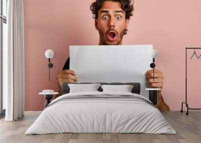  A man holding up an empty white sheet of paper with his mouth open, looking surprised and shocked isolated on a clear light background. Conceptual stock photo that could be used for advertising Wall mural