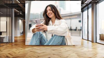 Bright and cheerful young woman with shopper on shoulder waiting public transport at station smiling beautifully while texting on phone Wall mural