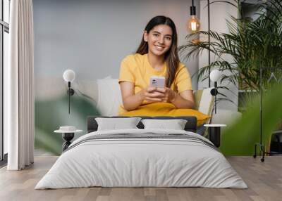 Cheerful female student with beautiful smile on cosy sofa at home holding a phone looking in the camera Wall mural