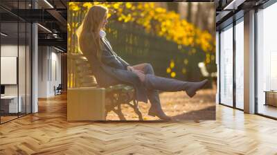 Beautiful girl with light brown hair is sitting on a bench in the alley of an autumn park. Attractive young woman in a gray coat and high boots. Fall season. Golden leaves. Amazing autumn mood. Wall mural