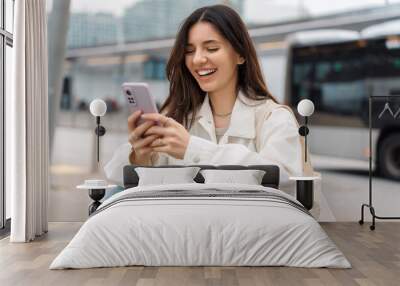 Waiting for the bus and for love. Bright cheerful young woman of Turkish mixed race in white casual clothes with a charming smile holding a phone in the street at public transportation stop Wall mural