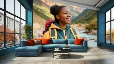 Young woman hiking, wearing a green and yellow jacket, stands by a mountain river, smiling confidently amidst a picturesque mountain range on a sunny day. Wall mural