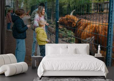 Family with child in zoo feeds buffalo. Happy family, young mother with three children, cute laughing toddler boy and a teen age girl and boy feeding buffalo during a trip to a city zoo on a hot Wall mural