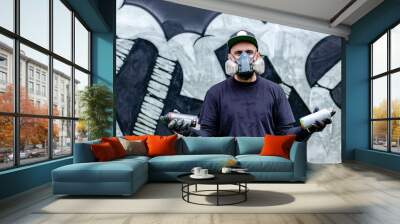 Graffiti artist posing in front of his drawing on the wall, with two aerosol spray paints in a can, wearing protective face mask / respirator with filters. Street art culture concept. Wall mural