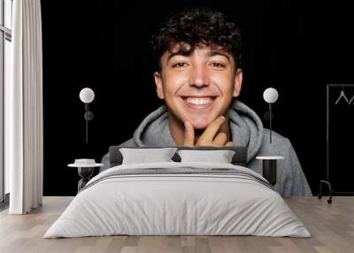 Portrait of young man about 25 years old with curly hair, holds hand on chin and smiles at camera. Isolated on black studio background Wall mural