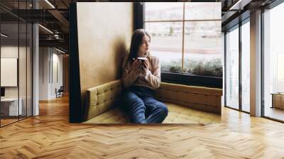 Cheerful young girl having breakfast at cozy home with big cup of coffee or tea holding in hands. Lifestyle, relaxation, natural beauty concept Wall mural