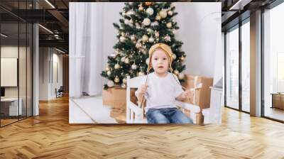 Infant baby boy in knitted hat playing at home in christmas evening. Holiday decorations, new year eve with colorful lights are on background Wall mural