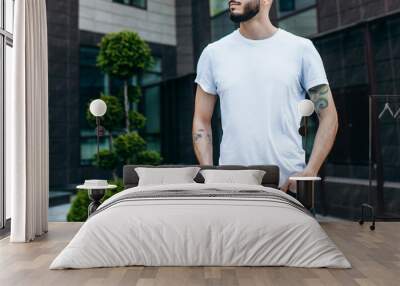 A young stylish man with a beard in a white T-shirt and glasses. Street photo Wall mural