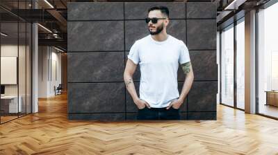 A young stylish man with a beard in a white T-shirt and glasses. Street photo Wall mural