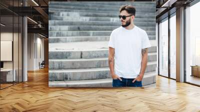 A young stylish man with a beard in a white T-shirt and glasses. Street photo Wall mural