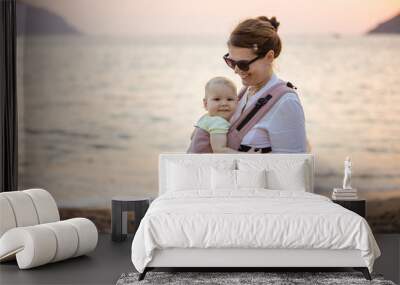 Cheerful Caucasian woman with little daughter in baby carrier on beach at sunset Wall mural