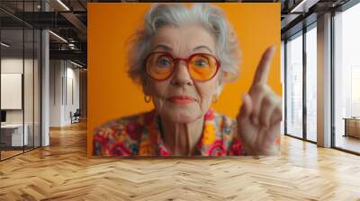 Elderly woman with orange glasses pointing upwards, vibrant orange background, expressing an idea or solution.
 Wall mural