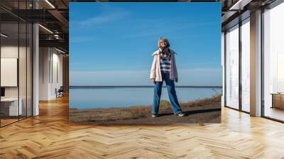 An adult woman in casual autumn clothes stands against the backdrop of a lake on a sunny autumn day. Wall mural