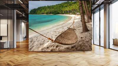 A wicker hammock hangs between two coconut trees against the backdrop of a tropical beach and blue sea. Wall mural