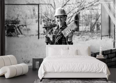 A young female bricklayer in an construction helmet with a construction hammer and a brick in her hands stands and smiles next to a brick wall black white photo Wall mural