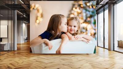 two funny little girls in a chair on the background of a Christmas tree. new year celebration with children. sisters on winter vacation. leisure with kids. traditional annual photo shoot. Wall mural