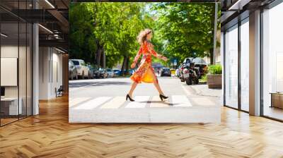 Young beautiful blonde woman in a red flower dress crosses the road at a crosswalk Wall mural