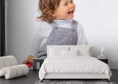 A cheerful laughing Caucasian toddler 2 years old with curly hair in a gray jumpsuit and a white turtleneck is playing with a car on the floor. Wall mural