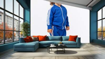 A cute curly-haired toddler of 1-2 years old stands sideways in a blue tracksuit and sneakers on a white one and looks away. Wall mural