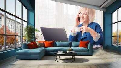 A cute smiling girl is talking on the phone and holding a cup of coffee or tea in her hand while working remotely from her home office. Wall mural