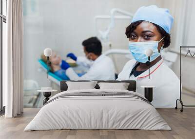 African dentist in mask looks into camera against the background of a doctor treats teeth Wall mural