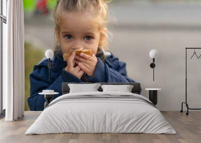 A beautiful little Caucasian girl with blond hair and eating bread eagerly with her hands looks at the camera with sad eyes Wall mural