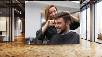 Handsome blue eyed man sitting in barber shop. Hairstylist Hairdresser Woman cutting his hair. Female barber. Wall mural